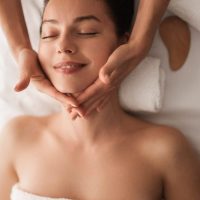 Top view of crop unrecognizable beautician doing face massage to delighted young female customer, lying on couch with closed eyes and smiling during spa treatment